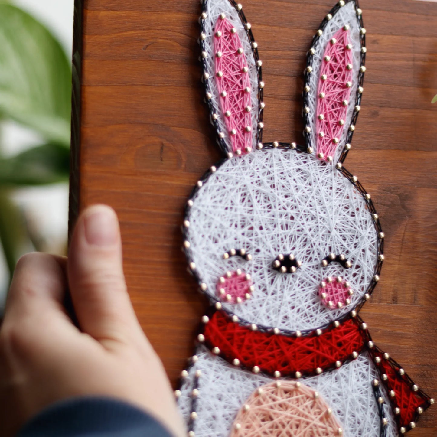 DIY string art kit featuring a cute bunny outlined with white, pink, and red thread on a rustic wooden board by GoodStrings - hand holding