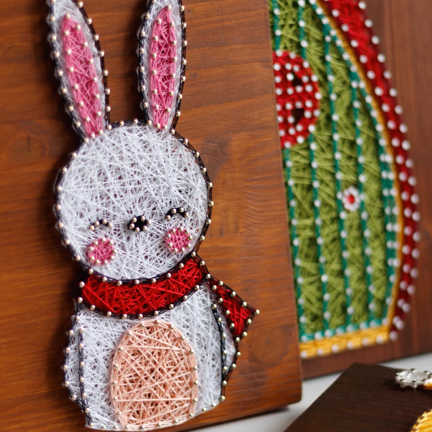 DIY string art kit featuring a cute bunny outlined with white, pink, and red thread on a rustic wooden board by GoodStrings - next to another string art sign