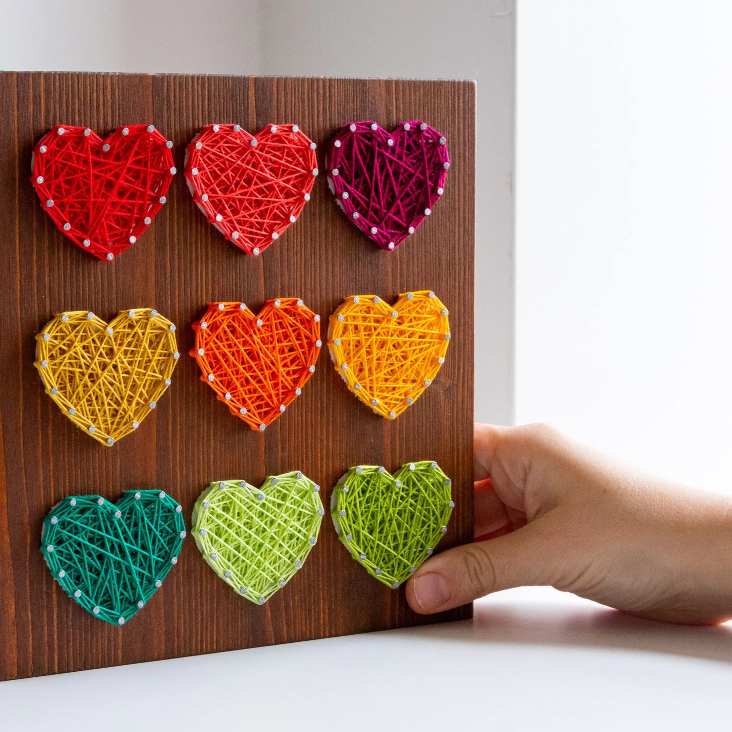 DIY string art kit with colorful hearts on a rustic wooden board by GoodStrings - hand touching for approximate scale