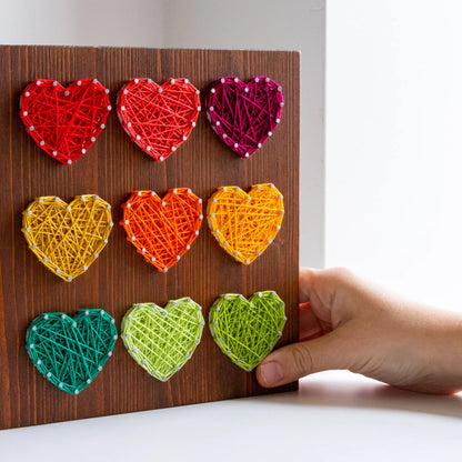 DIY string art kit with colorful hearts on a rustic wooden board by GoodStrings - hand touching for approximate scale