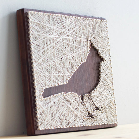 Side angle of a cardinal bird string art sign showing detailed string pattern on a dark wooden frame, resting on a wooden surface.