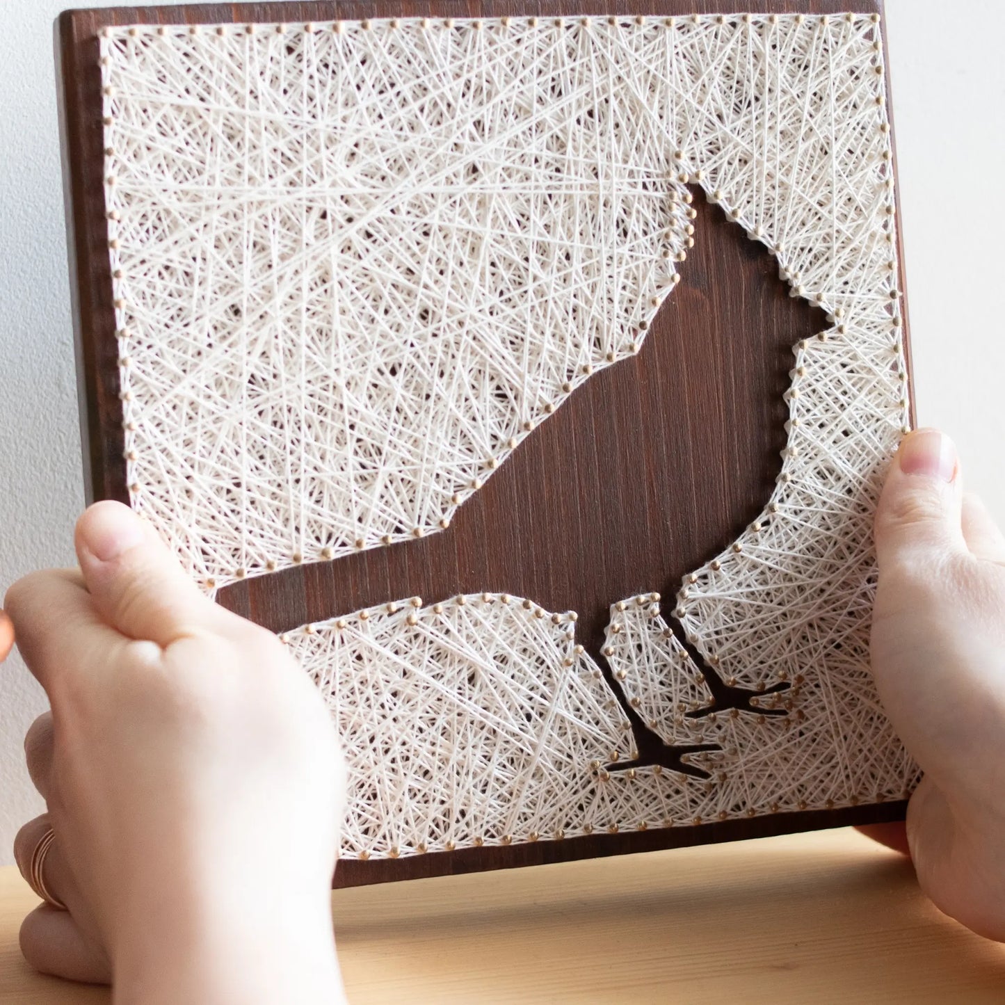 Side angle of a cardinal bird string art sign showing detailed string pattern on a dark wooden frame, resting on a wooden surface.