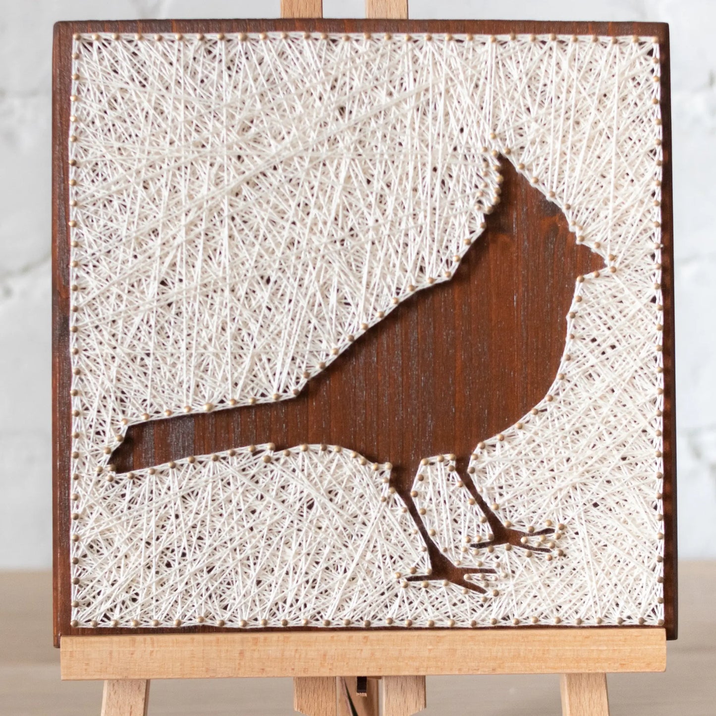 Side angle of a cardinal bird string art sign showing detailed string pattern on a dark wooden frame, resting on a wooden surface.