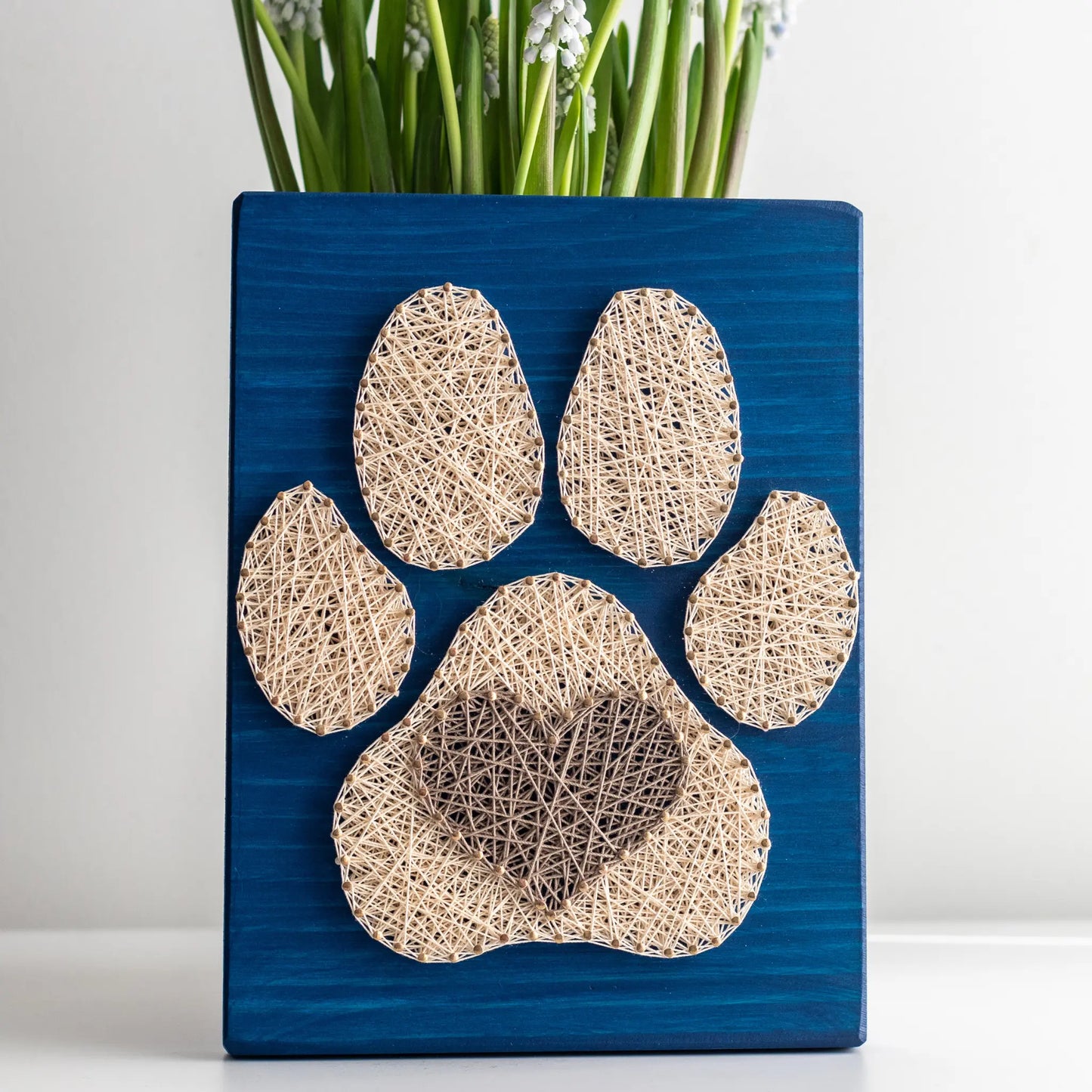  charming dog paw print string art sign made with white and dark brown string on a blue wooden background, featuring a heart shape in the center of the paw print.