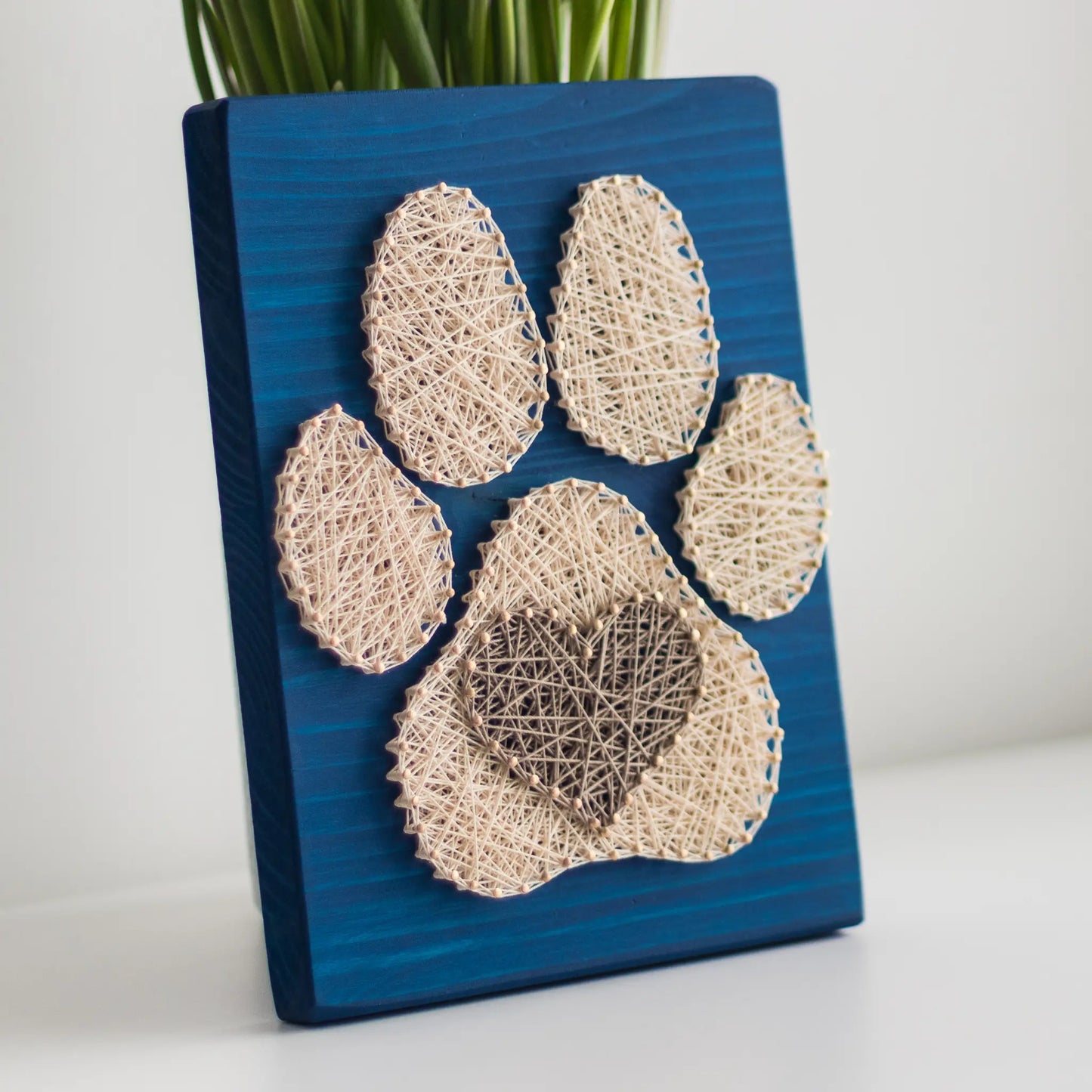  charming dog paw print string art sign made with white and dark brown string on a blue wooden background, featuring a heart shape in the center of the paw print.