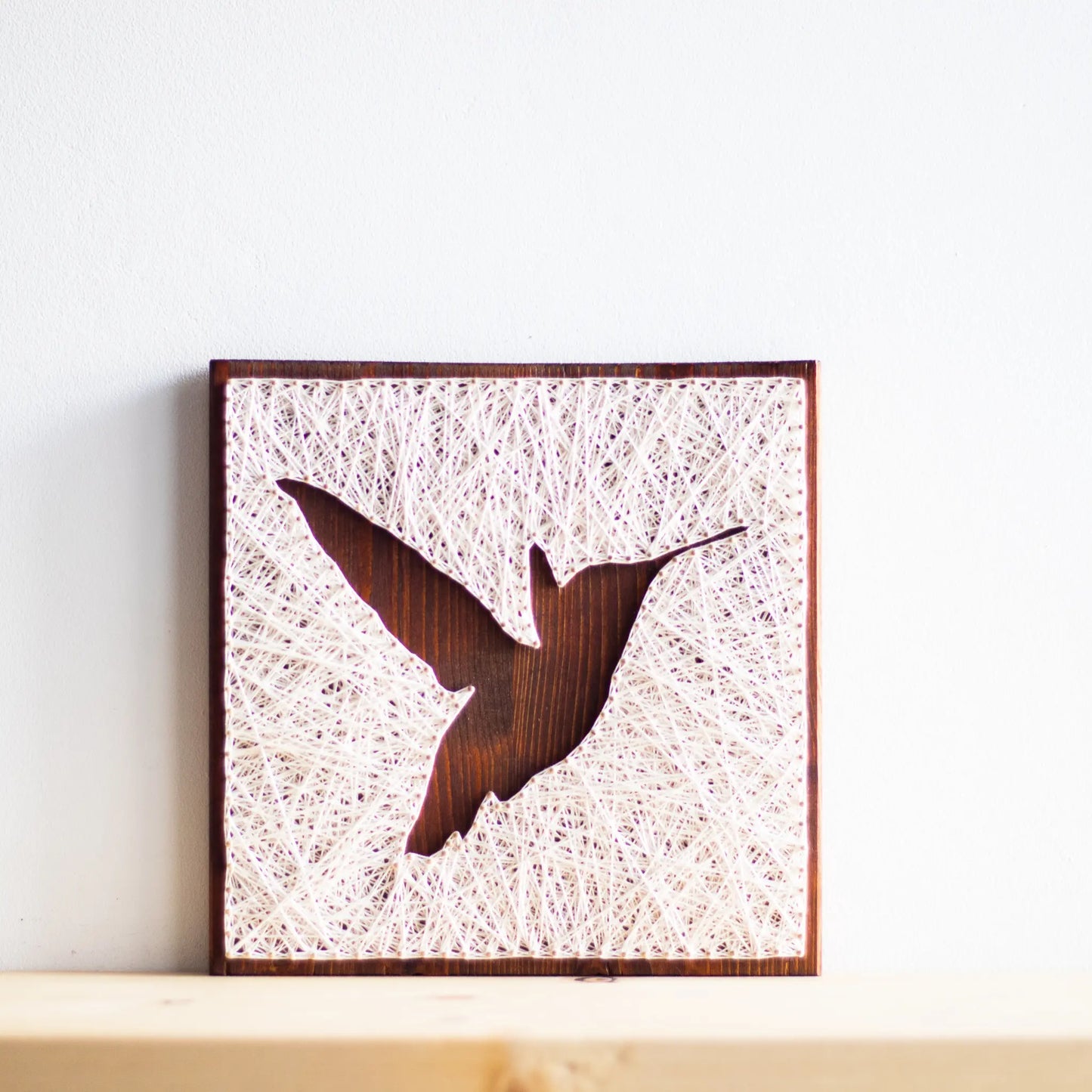 A beautiful hummingbird string art sign made with white string on a dark wooden background, depicting a hummingbird in mid-flight.