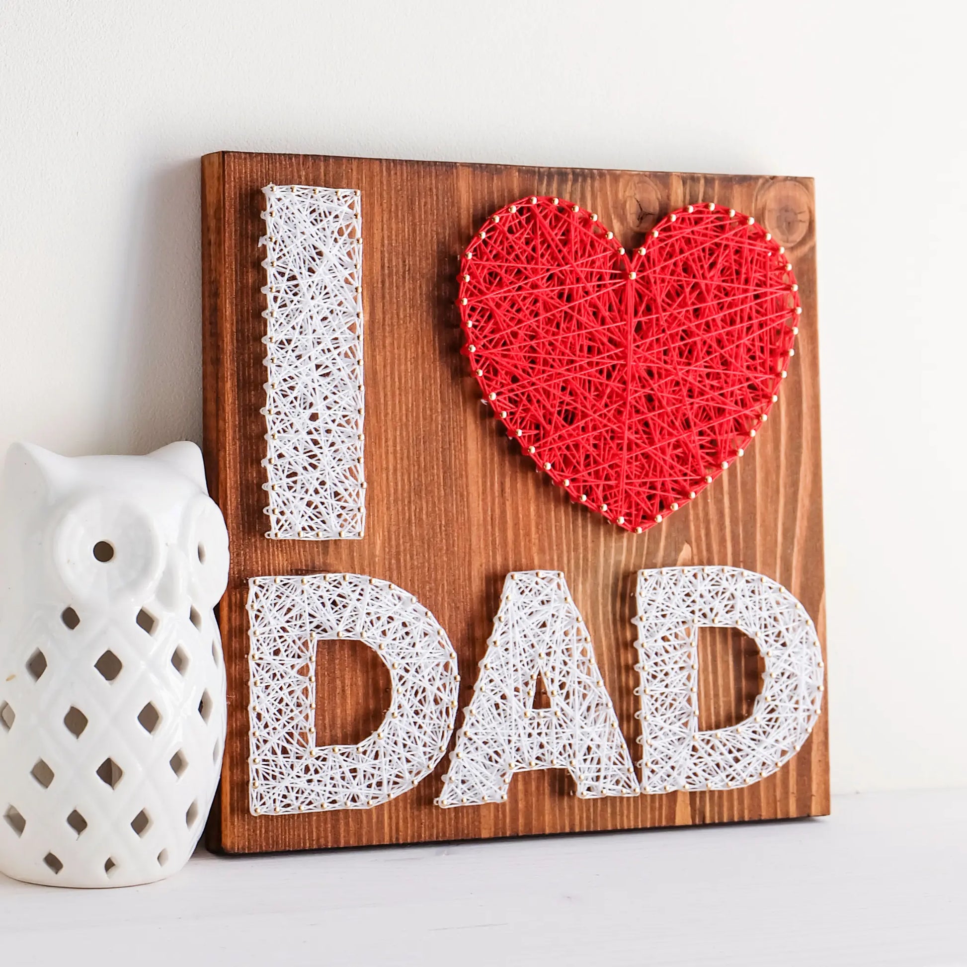 A heartfelt "I Love Dad" string art sign made with white and red string on a dark wooden background, featuring the words "I ♥ Dad" with a red heart.