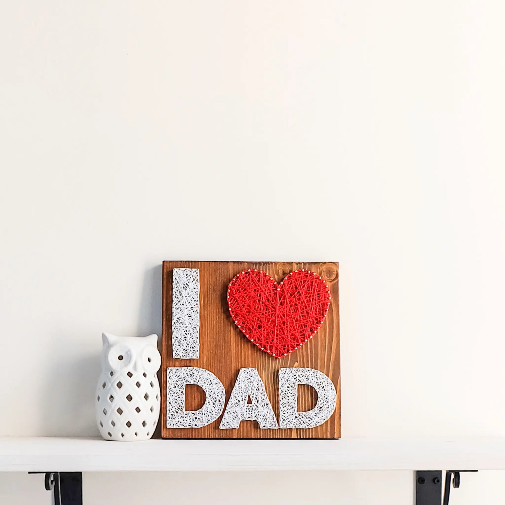 A heartfelt "I Love Dad" string art sign made with white and red string on a dark wooden background, featuring the words "I ♥ Dad" with a red heart.