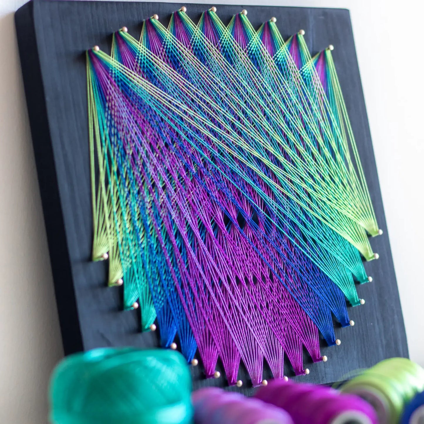 Geometric string art with green and purple threads on black base, part of craft kits for adults, highlighting intricate design.DIY geometric string art kit featuring a vibrant mandala design with multiple color options on a black wooden board by GoodStrings - green, purple and blue