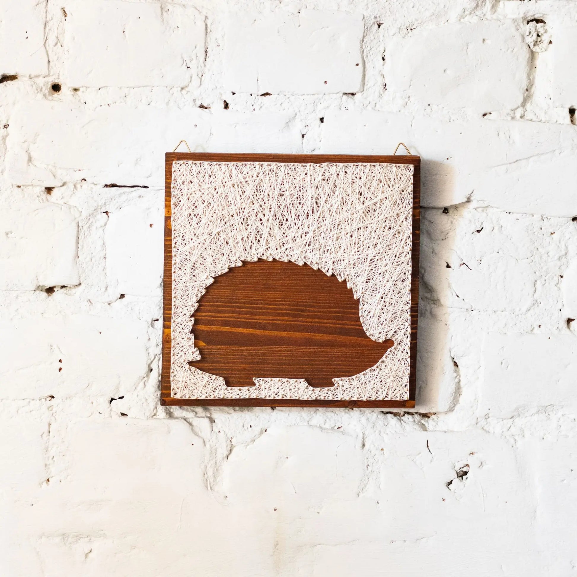 DIY string art kit featuring a hedgehog silhouette outlined with ivory thread on a rustic wooden board by GoodStrings - front view, white brick backgroubnd