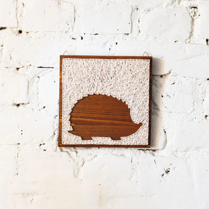 DIY string art kit featuring a hedgehog silhouette outlined with ivory thread on a rustic wooden board by GoodStrings - front view, white brick backgroubnd