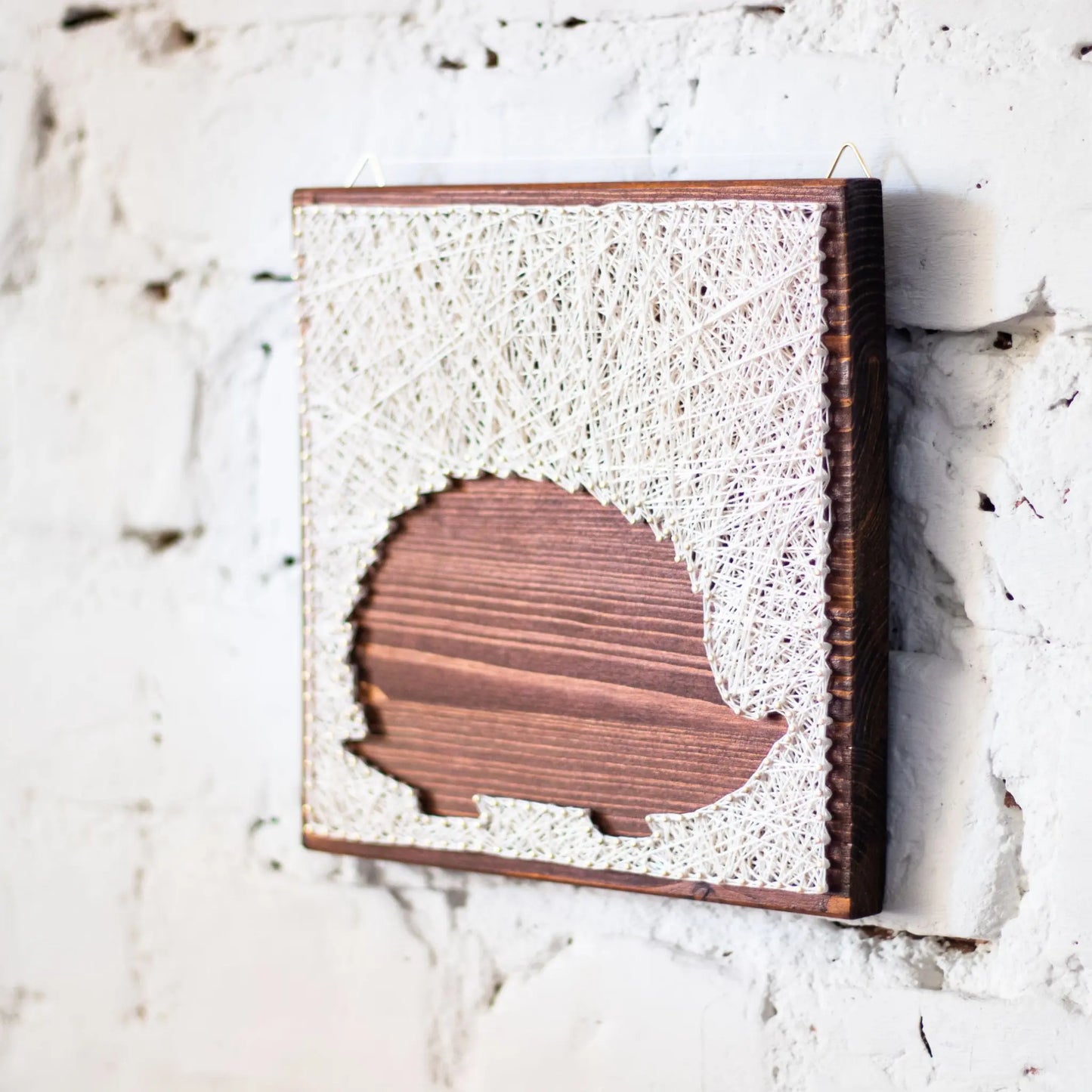 DIY string art kit featuring a hedgehog silhouette outlined with ivory thread on a rustic wooden board by GoodStrings - hanging on white brick wall - side view