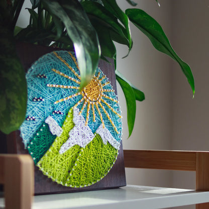 A piece of string art on a wooden board depicting a scenic mountain landscape. The landscape features green mountains with white snow-capped peaks, a bright yellow sun with rays, and a light blue sky with small clouds, all created using colorful strings and silver nails.