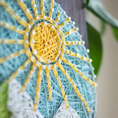 A piece of string art on a wooden board depicting a scenic mountain landscape. The landscape features green mountains with white snow-capped peaks, a bright yellow sun with rays, and a light blue sky with small clouds, all created using colorful strings and silver nails.