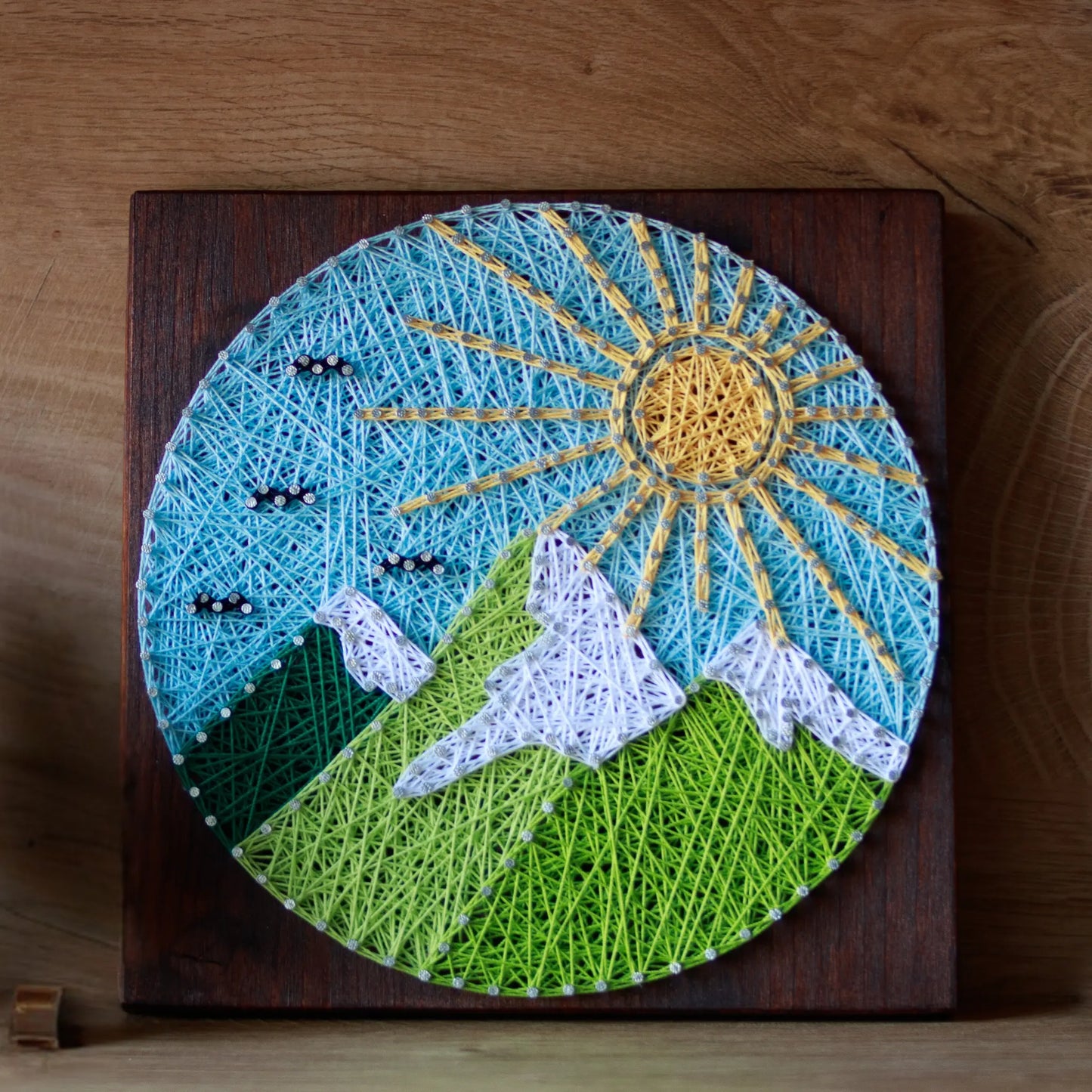 A piece of string art on a wooden board depicting a scenic mountain landscape. The landscape features green mountains with white snow-capped peaks, a bright yellow sun with rays, and a light blue sky with small clouds, all created using colorful strings and silver nails.