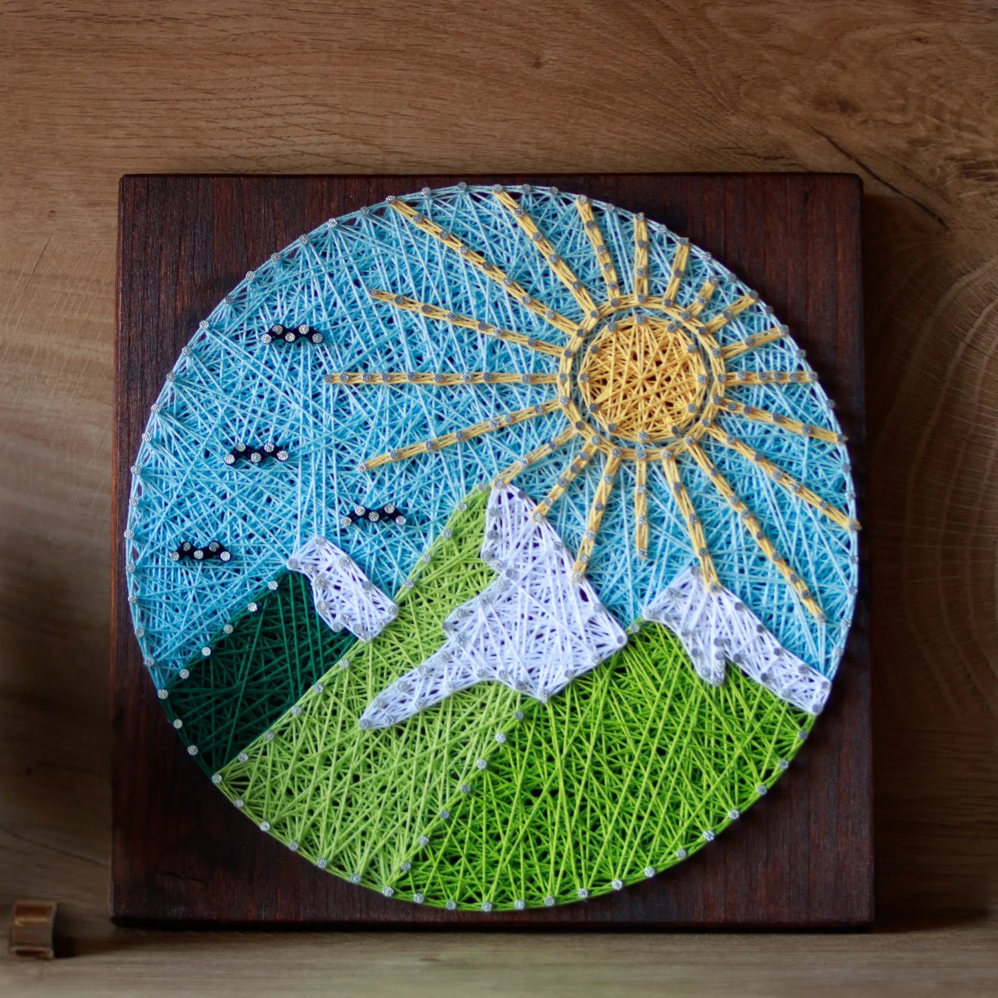 A piece of string art on a wooden board depicting a scenic mountain landscape. The landscape features green mountains with white snow-capped peaks, a bright yellow sun with rays, and a light blue sky with small clouds, all created using colorful strings and silver nails.