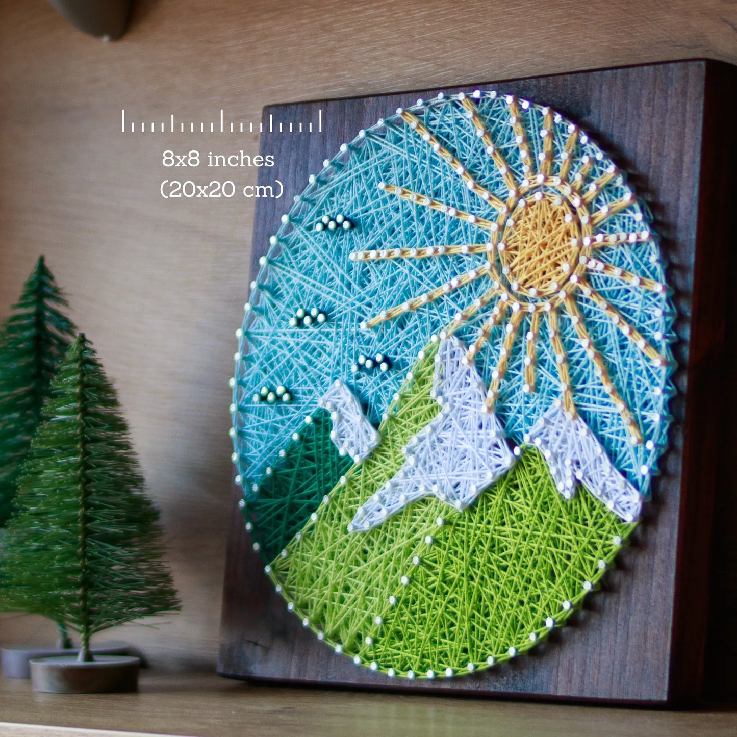 A piece of string art on a wooden board depicting a scenic mountain landscape. The landscape features green mountains with white snow-capped peaks, a bright yellow sun with rays, and a light blue sky with small clouds, all created using colorful strings and silver nails.