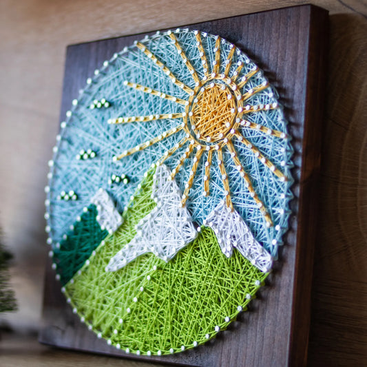 A piece of string art on a wooden board depicting a scenic mountain landscape. The landscape features green mountains with white snow-capped peaks, a bright yellow sun with rays, and a light blue sky with small clouds, all created using colorful strings and silver pins.