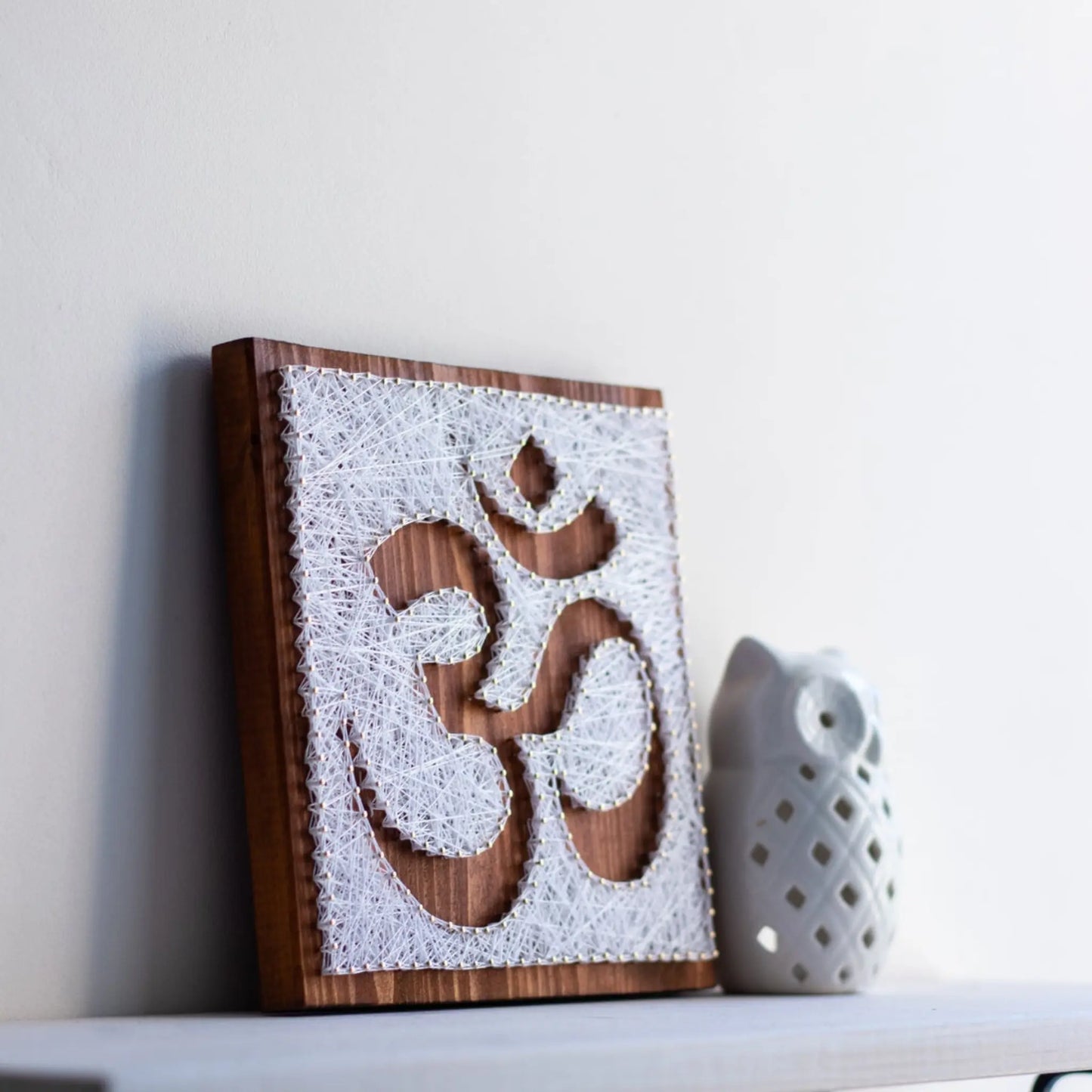 DIY string art kit featuring an Om symbol outlined with white thread on a rustic wooden board by GoodStrings - side view on shelf