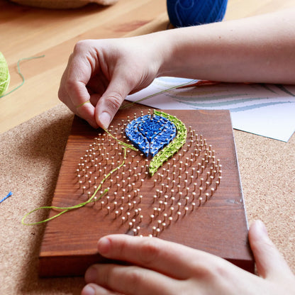 DIY string art kit featuring a peacock feather design with vibrant blue, green, and lime threads on a rustic wooden board by GoodStrings - being made