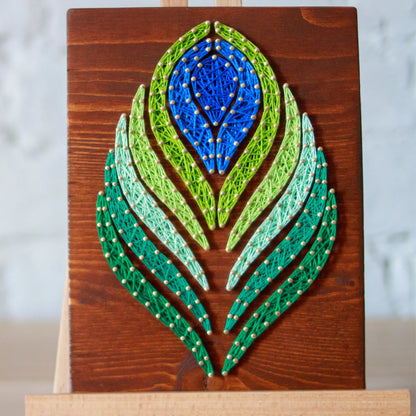 DIY string art kit featuring a peacock feather design with vibrant blue, green, and lime threads on a rustic wooden board by GoodStrings - front view