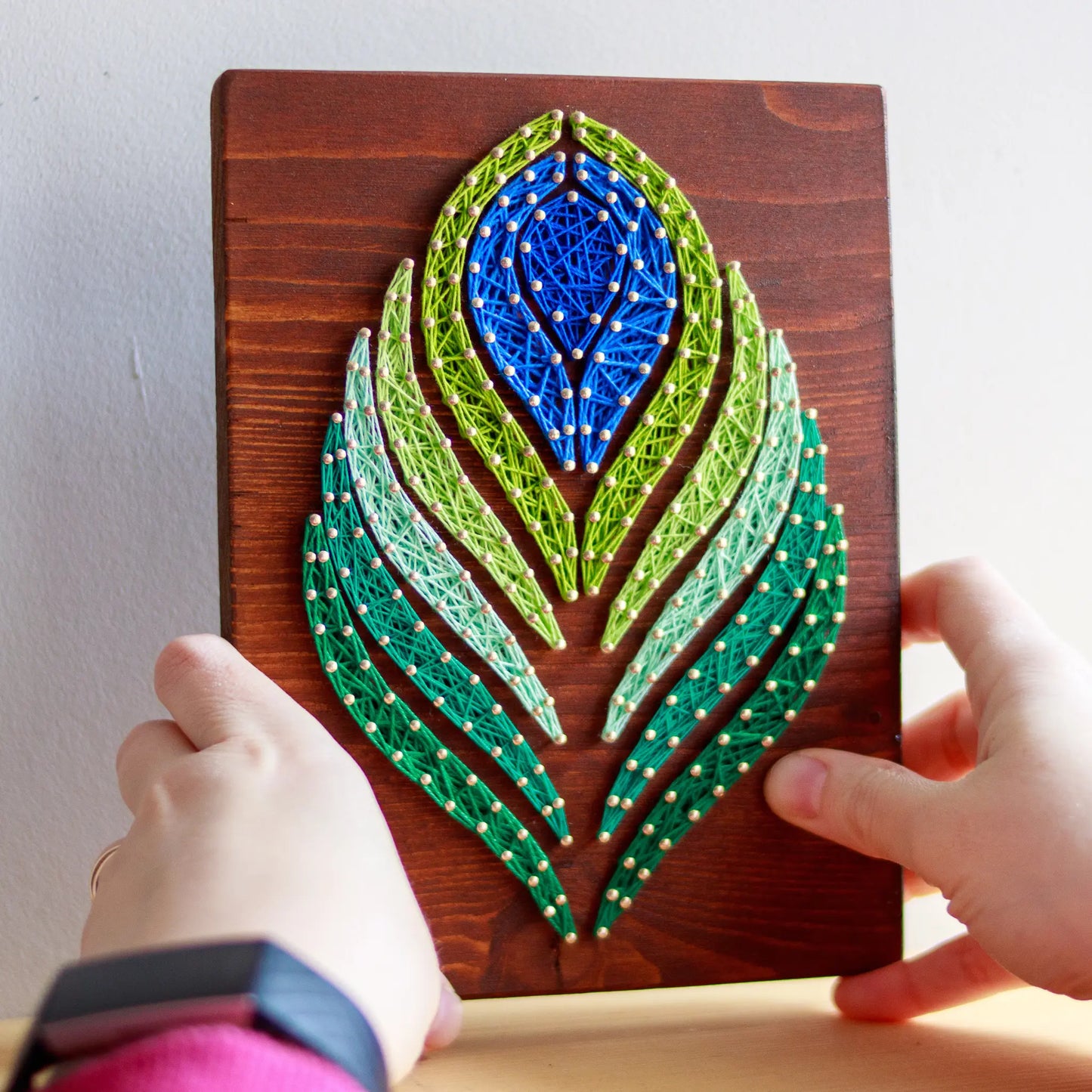 DIY string art kit featuring a peacock feather design with vibrant blue, green, and lime threads on a rustic wooden board by GoodStrings - hands holding