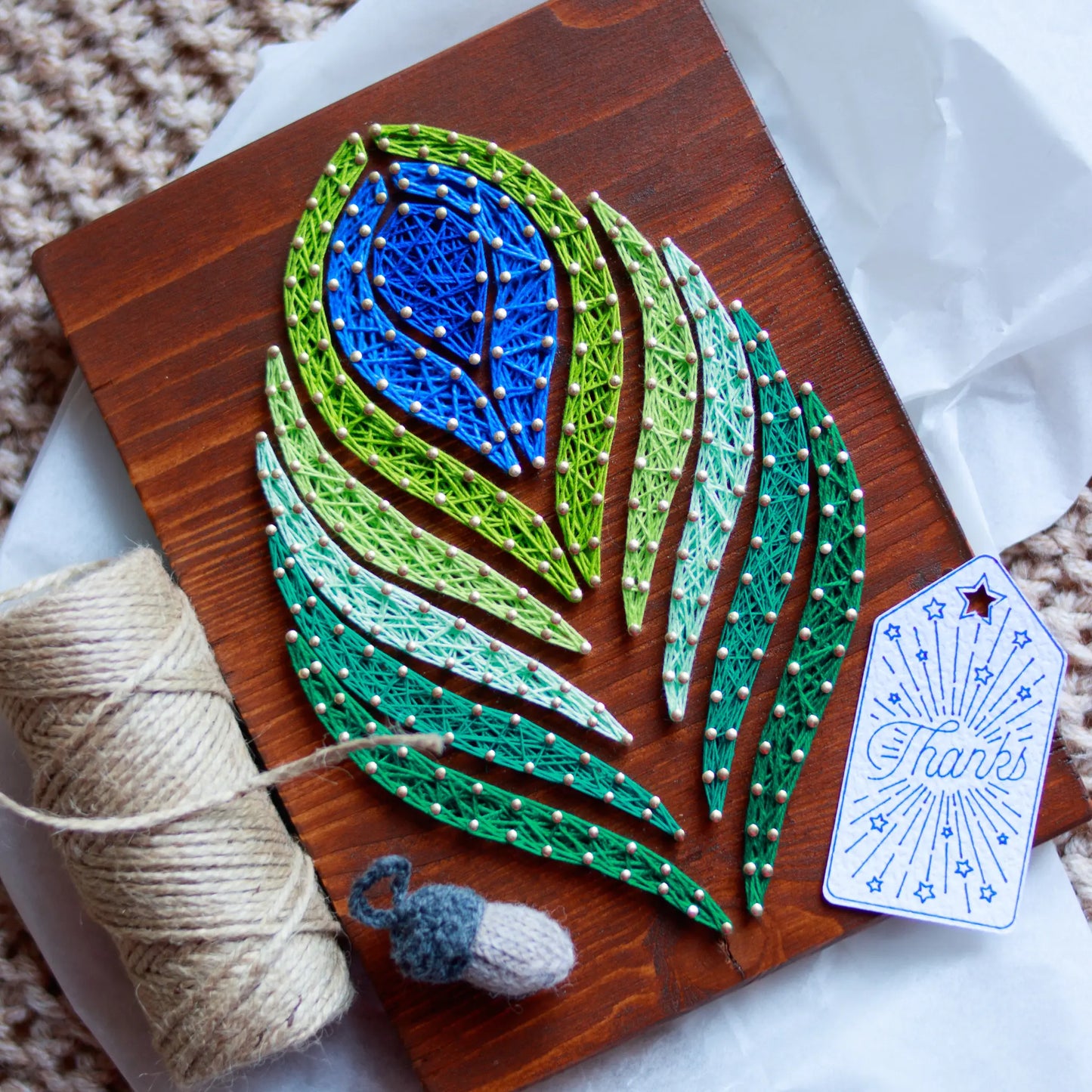 DIY string art kit featuring a peacock feather design with vibrant blue, green, and lime threads on a rustic wooden board by GoodStrings - top view