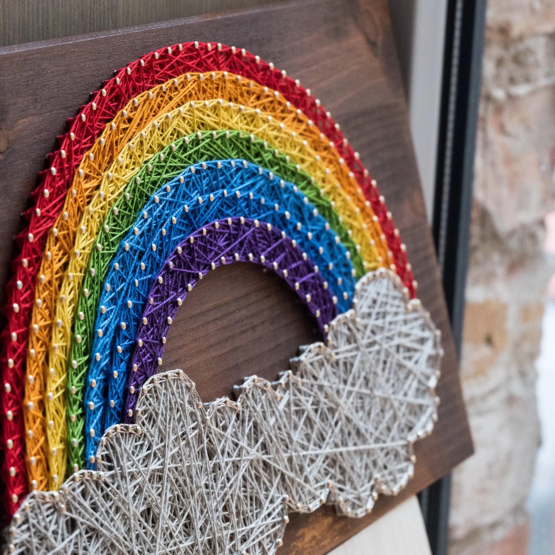 DIY string art kit featuring a vibrant rainbow arching over fluffy clouds, crafted with colorful threads on a dark wooden board, designed by GoodStrings