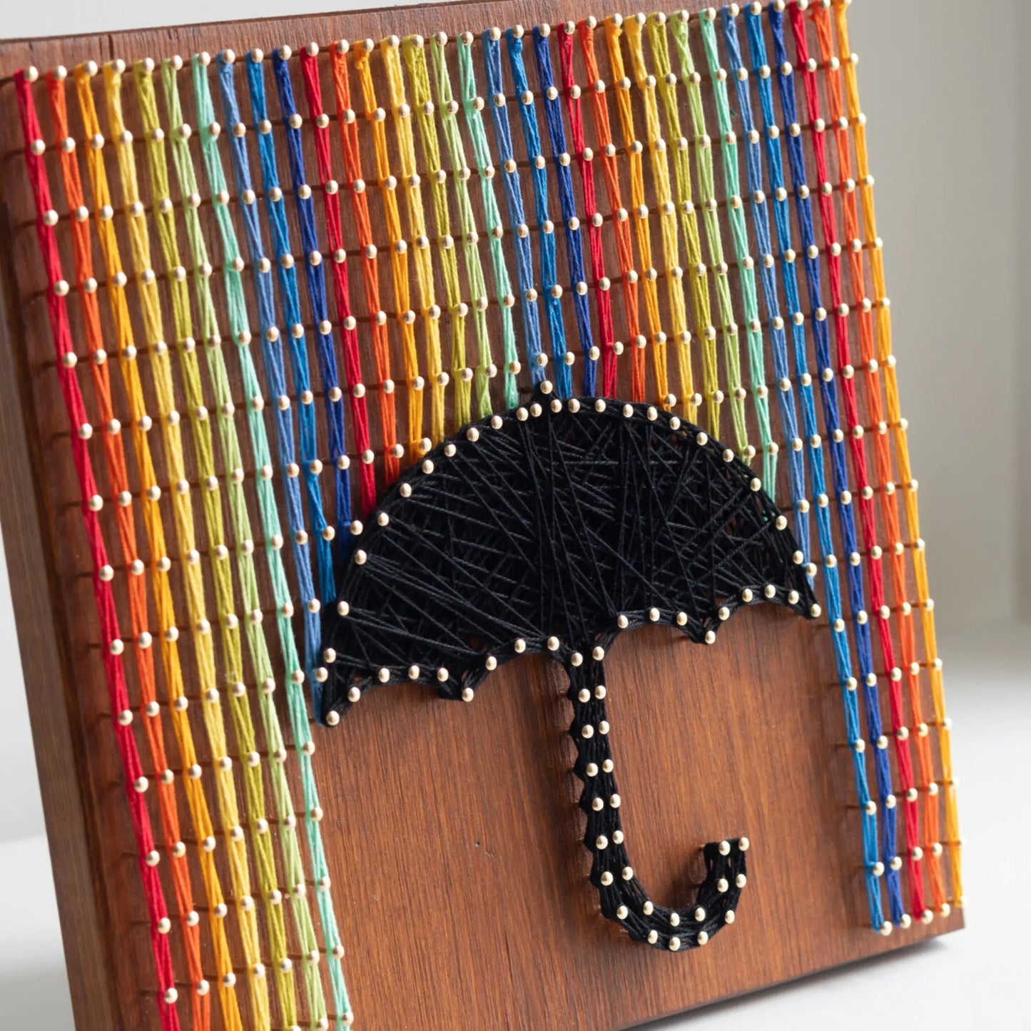 A piece of string art on a wooden board depicting a black umbrella. The umbrella is created using black string and gold pins. Above the umbrella, vertical rows of colorful strings in red, yellow, blue, orange, and green are arranged, representing rain falling from the top of the board.