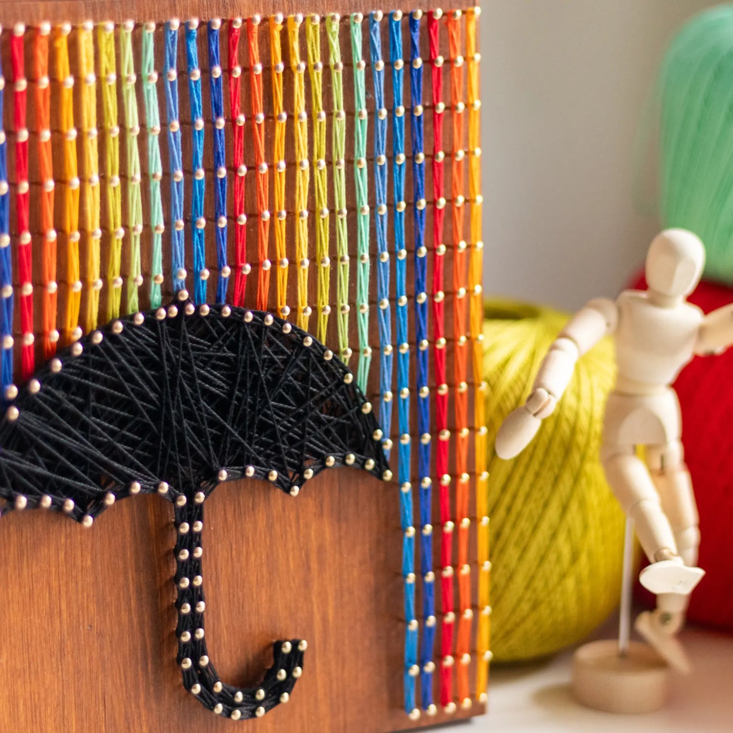 A piece of string art on a wooden board depicting a black umbrella. The umbrella is created using black string and gold pins. Above the umbrella, vertical rows of colorful strings in red, yellow, blue, orange, and green are arranged, representing rain falling from the top of the board.