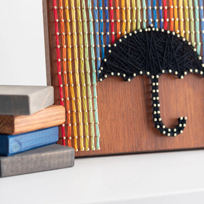 A piece of string art on a wooden board depicting a black umbrella. The umbrella is created using black string and gold pins. Above the umbrella, vertical rows of colorful strings in red, yellow, blue, orange, and green are arranged, representing rain falling from the top of the board.