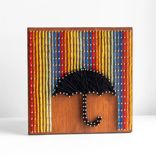 A piece of string art on a wooden board depicting a black umbrella. The umbrella is created using black string and gold pins. Above the umbrella, vertical rows of colorful strings in red, yellow, blue, orange, and green are arranged, representing rain falling from the top of the board.