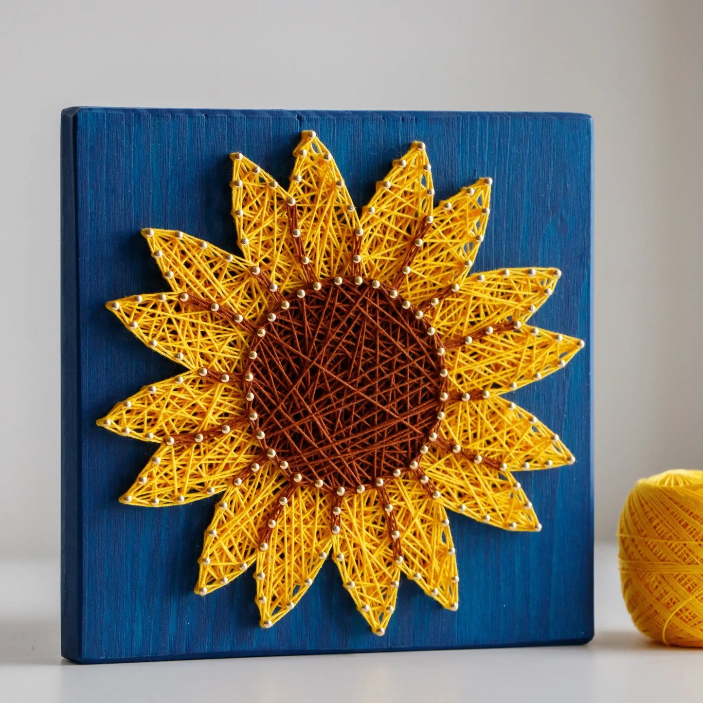 DIY string art kit featuring a bright yellow sunflower with a brown center created with threads on a dark blue wooden board by GoodStrings