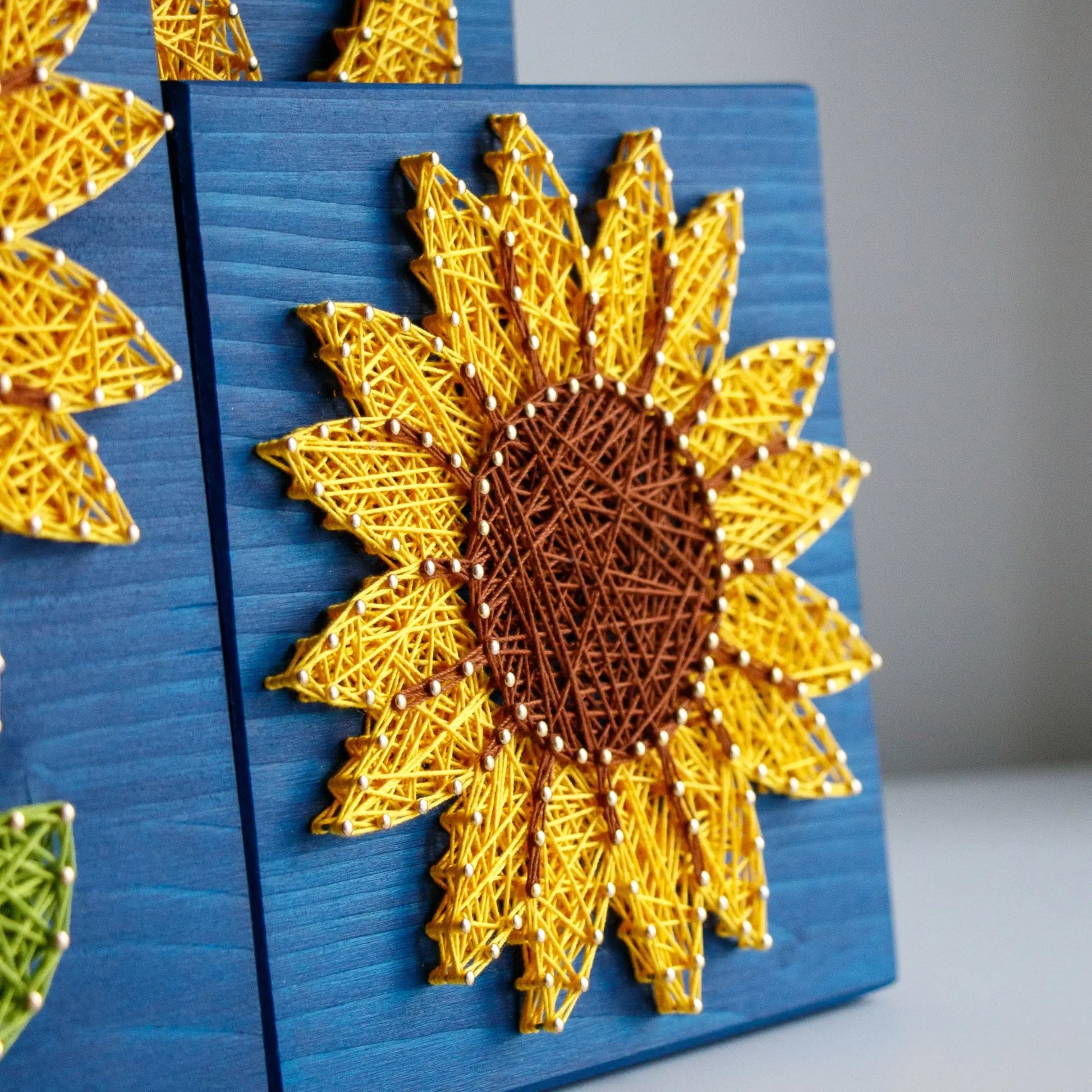 DIY string art kit featuring a bright yellow sunflower with a brown center created with threads on a dark blue wooden board by GoodStrings