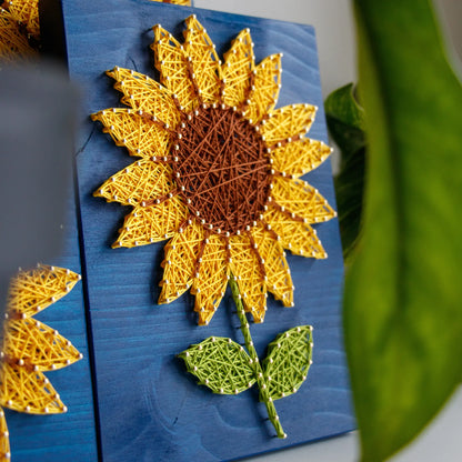 DIY string art kit featuring a bright yellow sunflower with a brown center and green leaves created with threads on a dark blue wooden board by GoodStrings