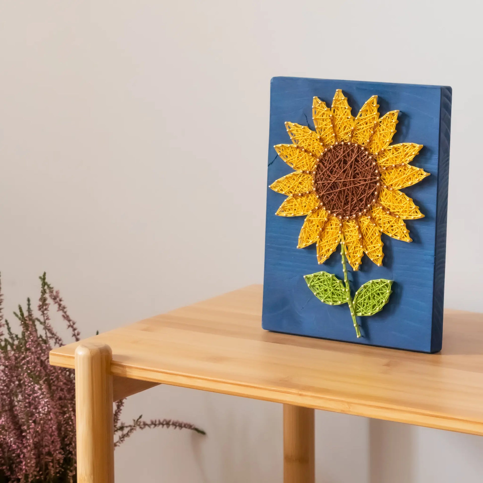 DIY string art kit featuring a bright yellow sunflower with a brown center and green leaves created with threads on a dark blue wooden board by GoodStrings