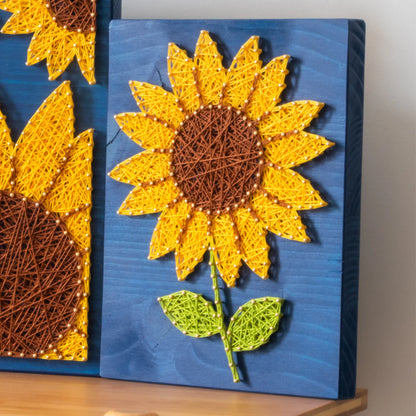 DIY string art kit featuring a bright yellow sunflower with a brown center and green leaves created with threads on a dark blue wooden board by GoodStrings