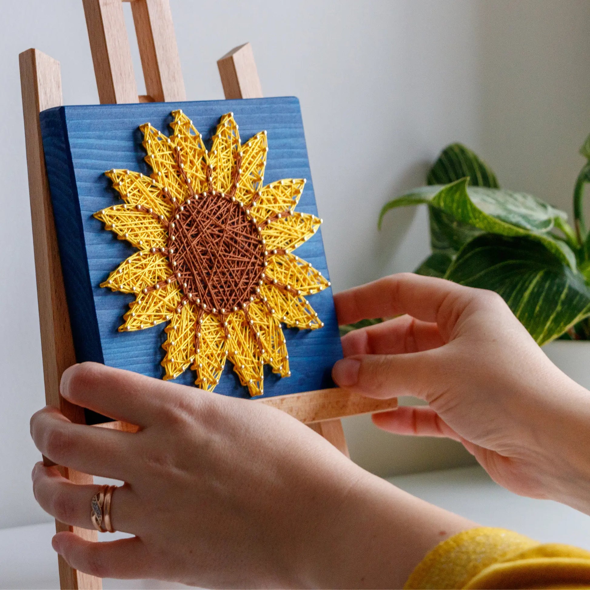 DIY string art kit featuring a bright yellow sunflower with a brown center and green leaves created with threads on a dark blue wooden board by GoodStrings