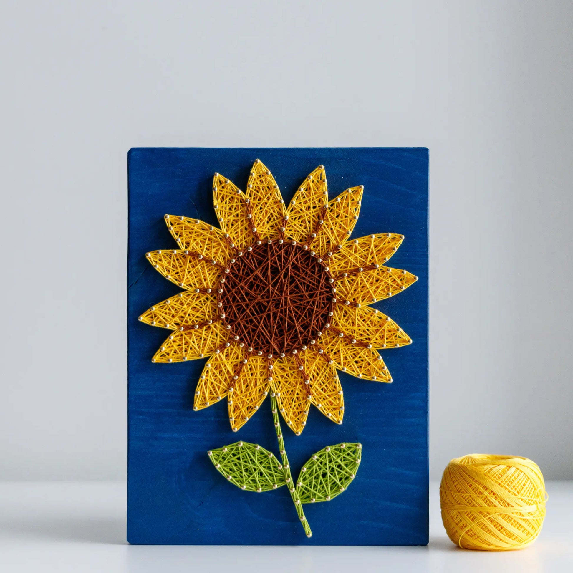 DIY string art kit featuring a bright yellow sunflower with a brown center and green leaves created with threads on a dark blue wooden board by GoodStrings