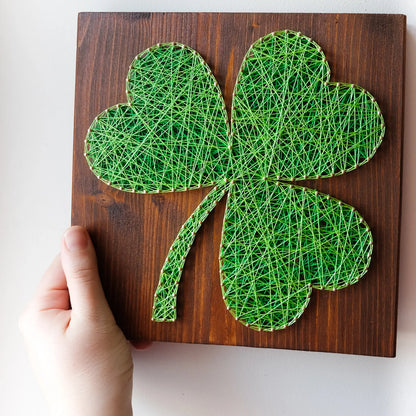 DIY string art kit featuring a three-leaf clover outlined with green thread on a rustic wooden board by GoodStrings - hand holding