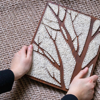 Close-up of the string art process, showing hands weaving ivory thread to form a tree silhouette on a wooden board, part of a craft kit for adults.