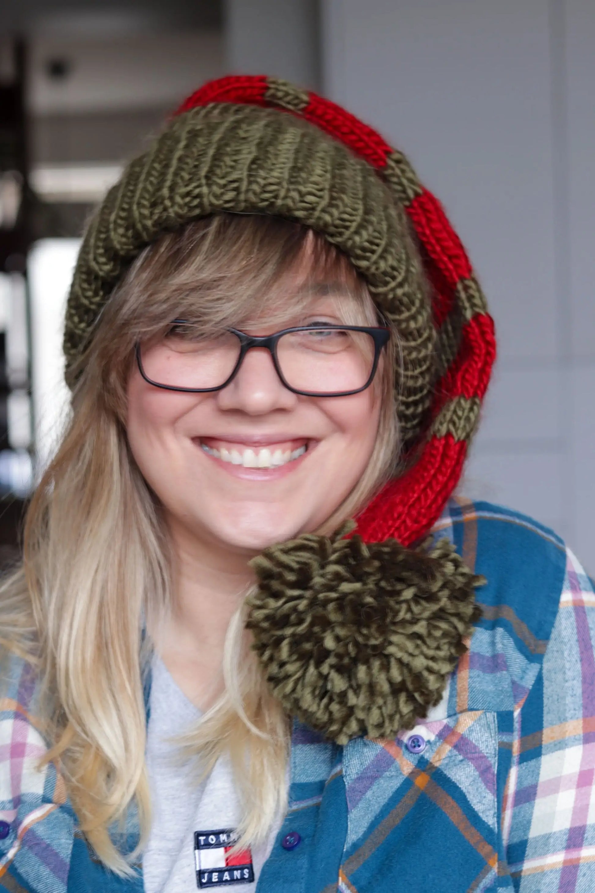 Smiling woman wearing a green and red striped knitted Santa hat with a large green pom-pom, styled casually with glasses and a plaid shirt.