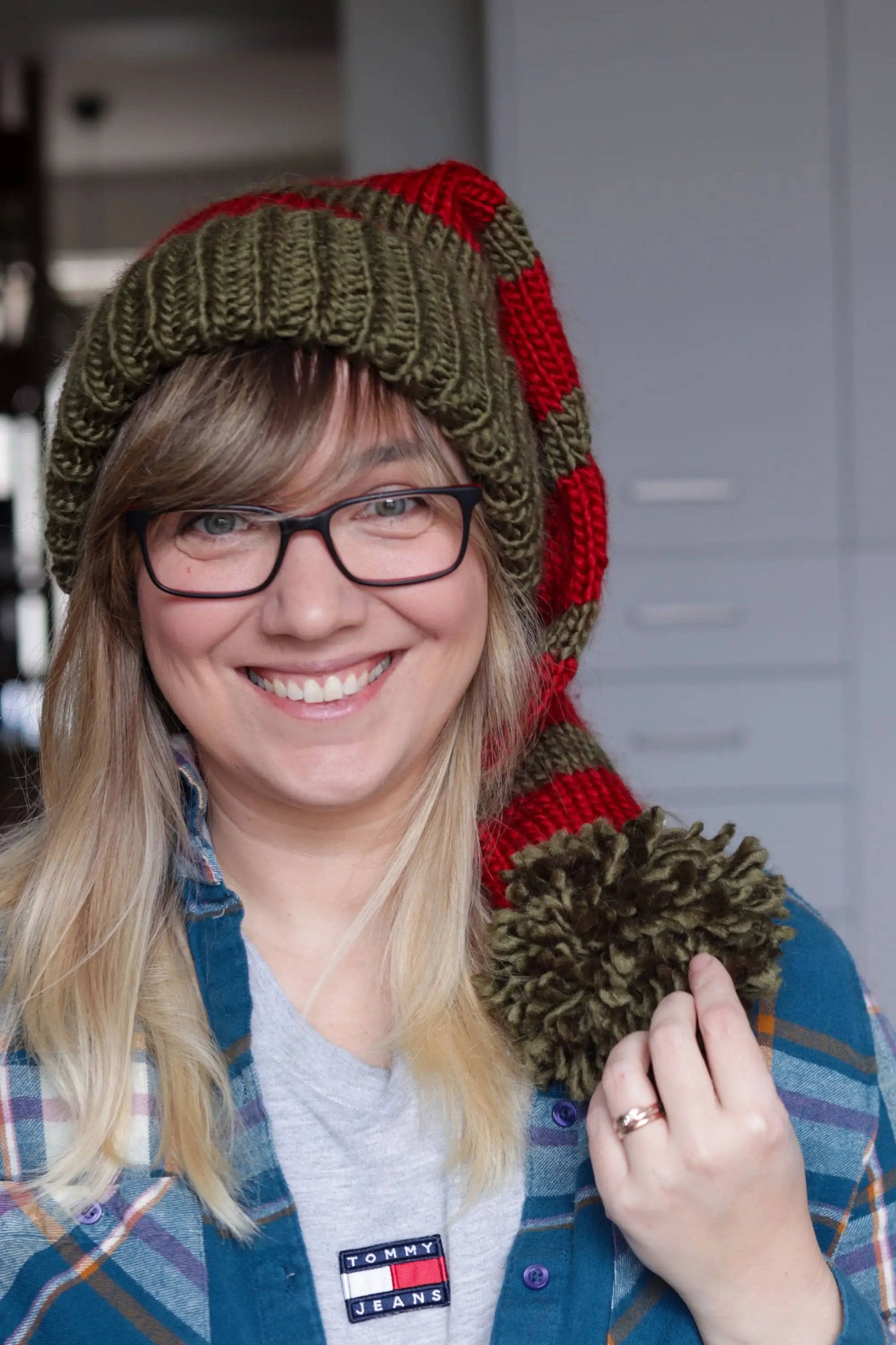 Woman smiling warmly while wearing a green and red knitted Santa hat with a big green pom-pom, holding the pom-pom in her hand, paired with a plaid shirt.