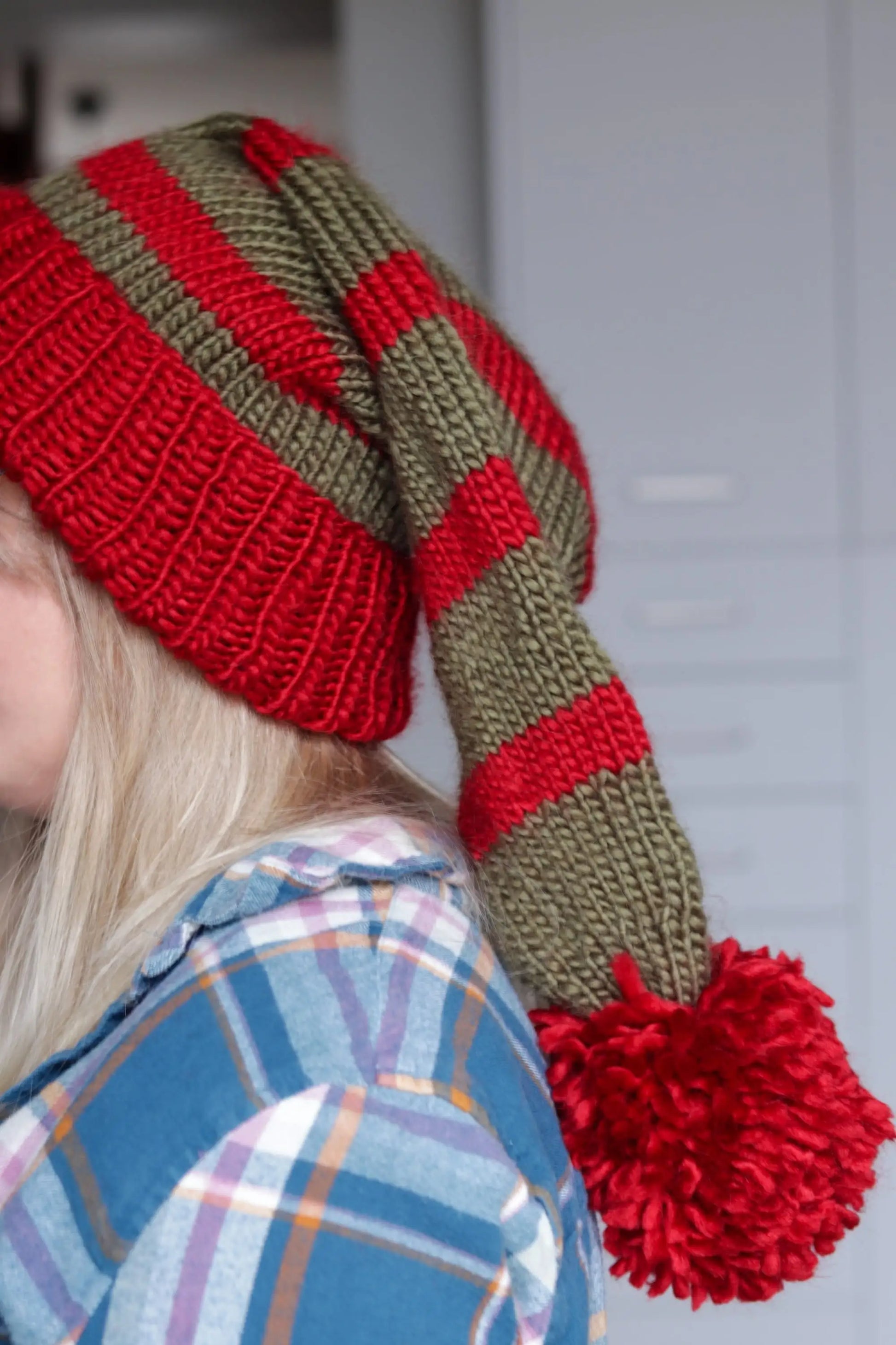 Side profile of a green and red striped knitted Santa hat with a long tail and a fluffy red pom-pom, worn by a woman in a plaid shirt.