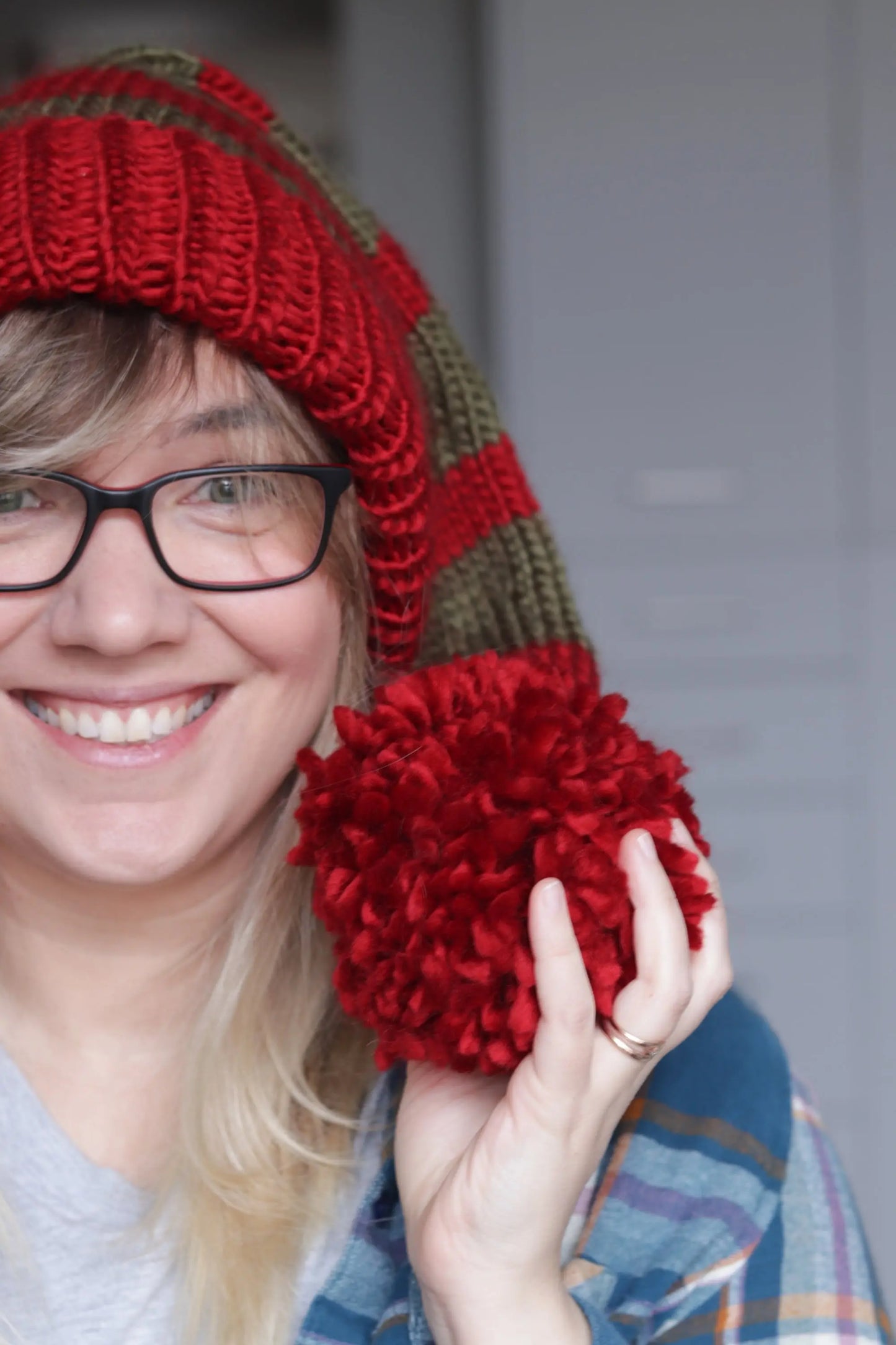 Woman smiling and showcasing the red pom-pom of her green and red striped knitted Santa hat, exuding festive charm.