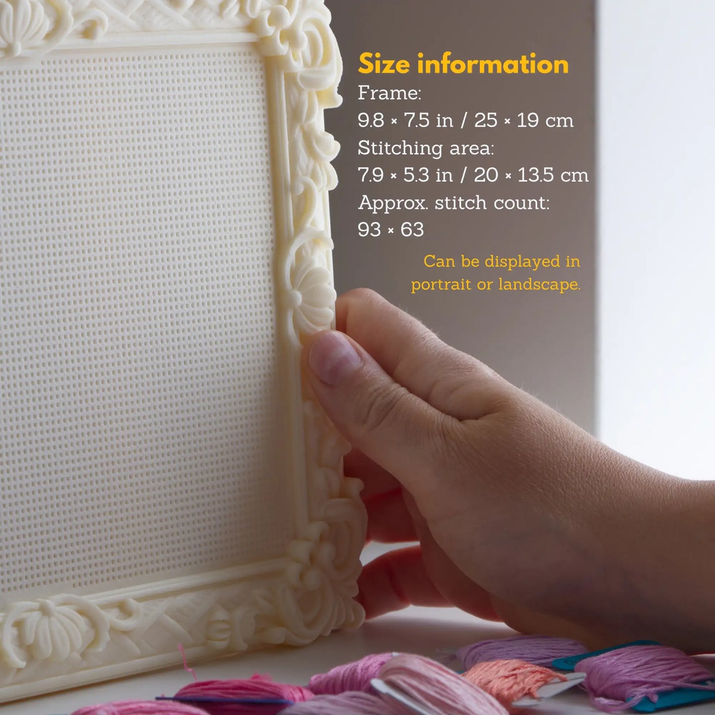 Close-up of a hand holding an ornate cream-colored embroidery frame with a white stitched canvas insert. Yellow text on the right lists size information for the frame and stitching area.