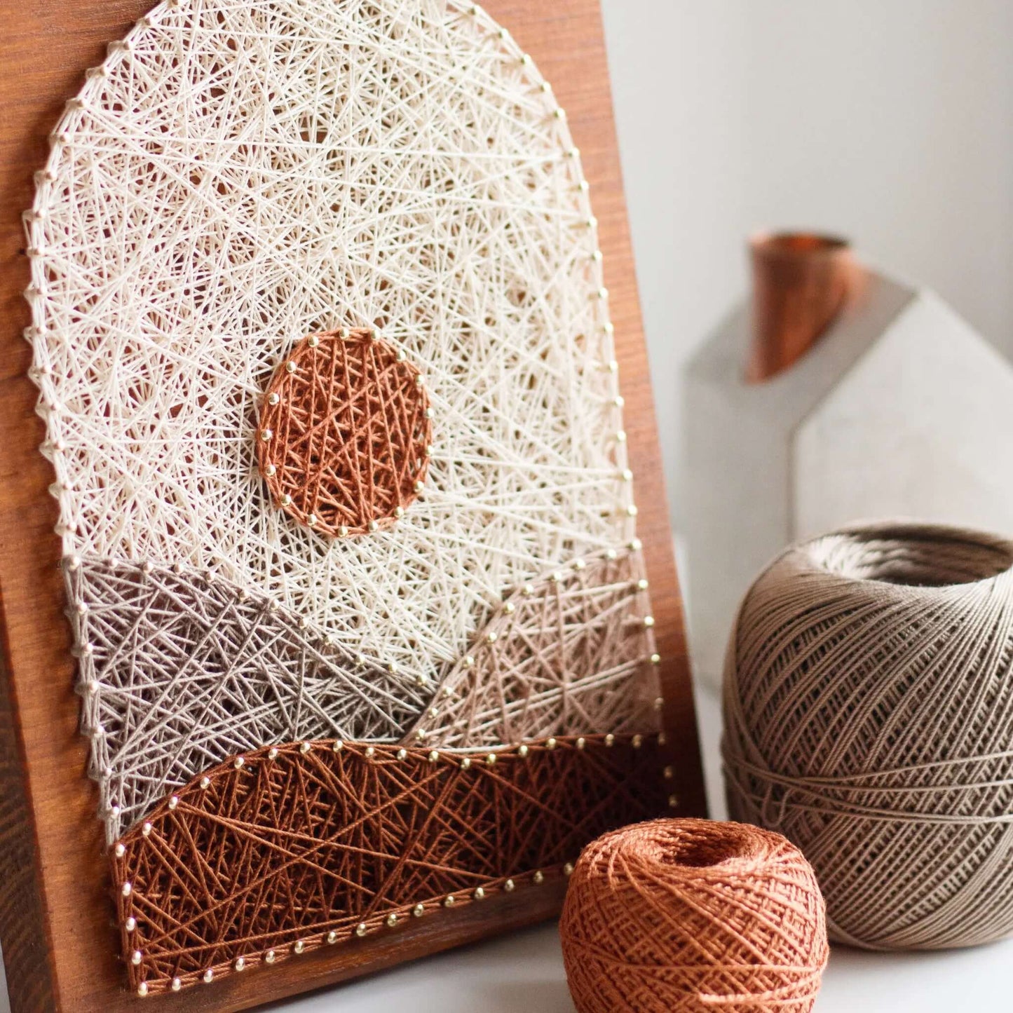 A piece of string art on a wooden board depicting a minimalist landscape. The design features a brown sun near the top center and abstract geometric hills in shades of brown, beige, and tan. The hills and sun are created using various shades of string and gold pins, giving a textured appearance to the artwork.