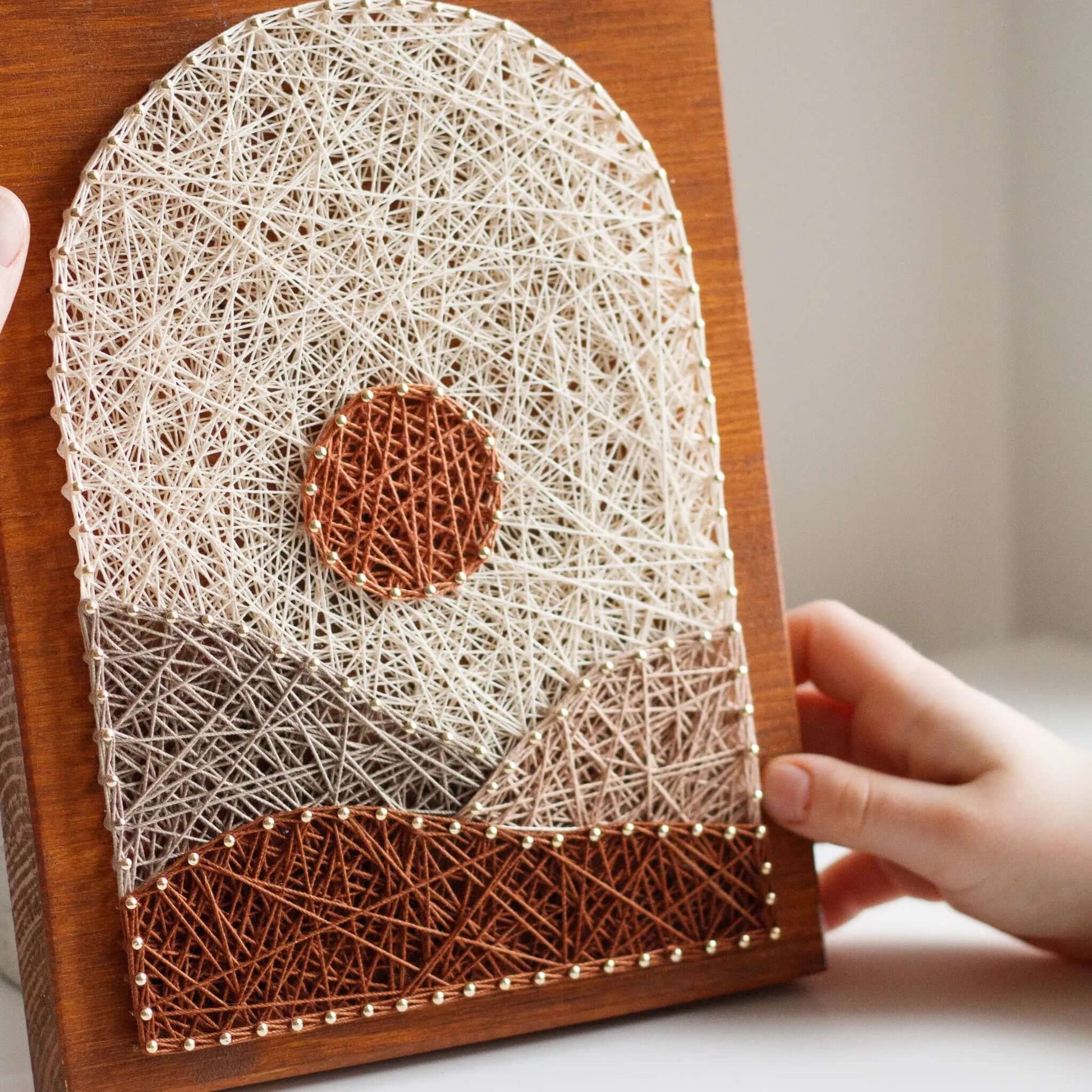 A piece of string art on a wooden board depicting a minimalist landscape. The design features a brown sun near the top center and abstract geometric hills in shades of brown, beige, and tan. The hills and sun are created using various shades of string and gold pins, giving a textured appearance to the artwork.