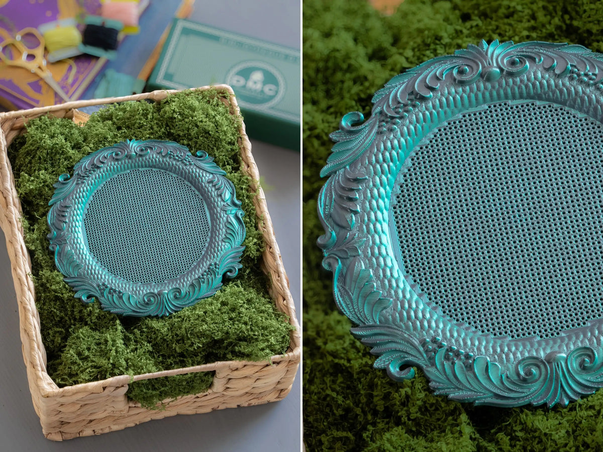 Round 3D printed cross stitch frame in Nightshade green, ornate feather-scroll border with a blank stitching grid, in a moss-filled basket.