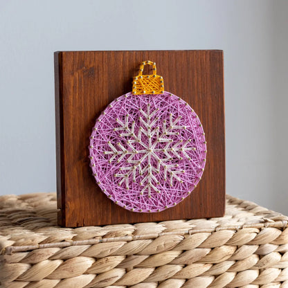 Decorative string art ornament on a wooden block with a purple ball and gold top, placed on a woven surface.